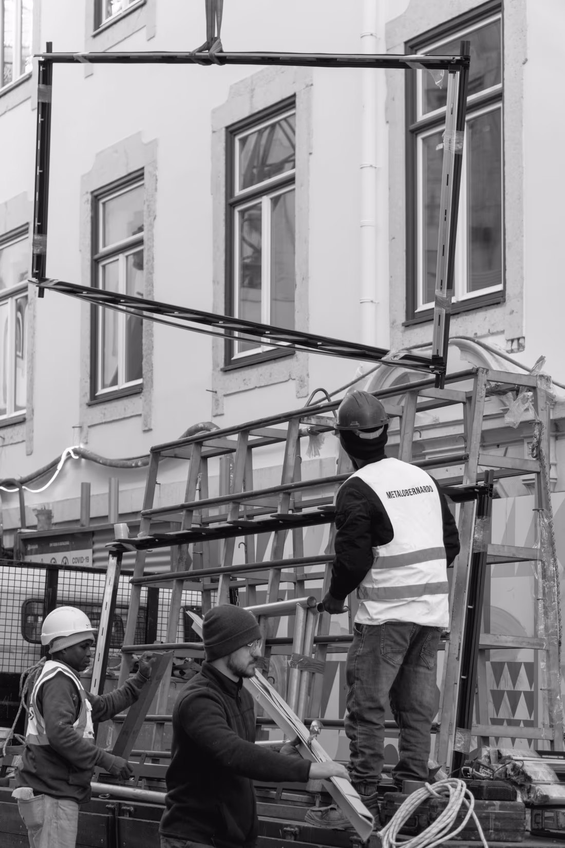 Glazed window units stacked upright in a padded transport rack on a job site, with crew members handling a frame