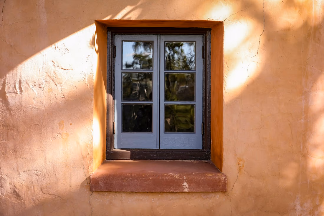 A small window set deep in a thick stucco-rendered masonry wall, with the render carried into the reveal and a projecting sill below
